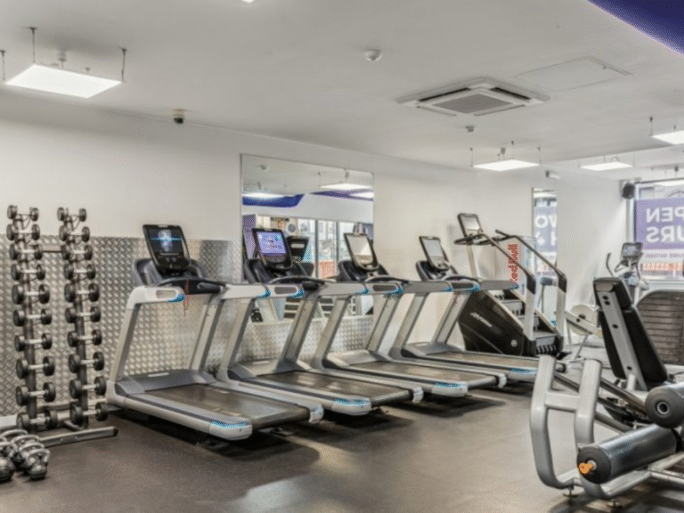 A modern gym with several treadmills, an elliptical machine, and rows of dumbbells on racks against a mirrored wall. The space is bright and clean with exercise equipment neatly arranged.