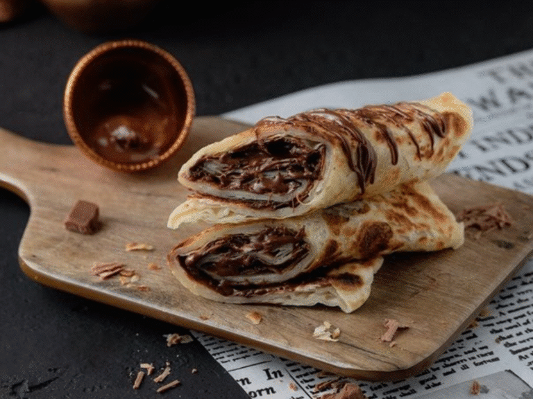Two stuffed crepes filled with chocolate spread are stacked on a wooden board, drizzled with more chocolate, with a small cup and chocolate shavings nearby on a newspaper background.