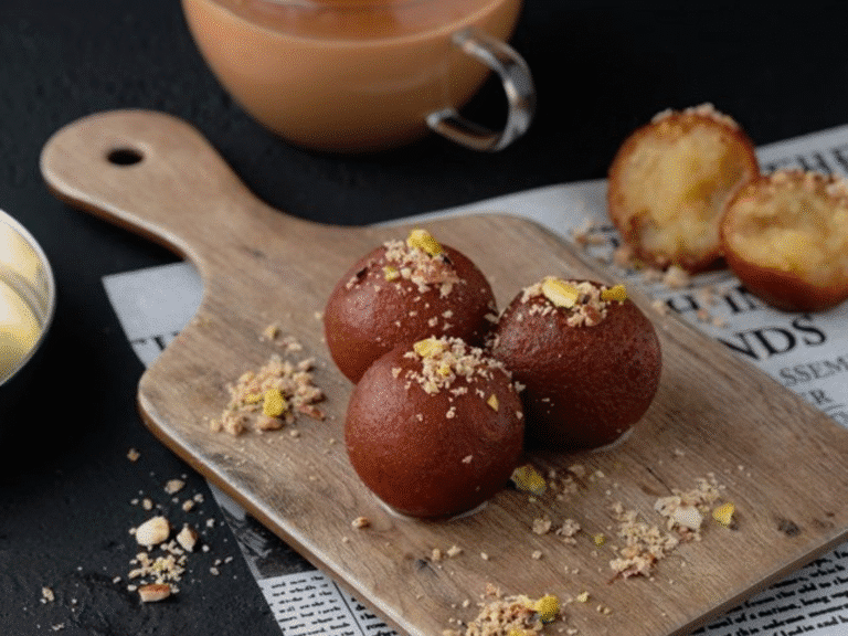 Three pieces of gulab jamun topped with crushed nuts are placed on a wooden board, with a tea cup and a halved gulab jamun in the background, all set on a newspaper over a dark surface.
