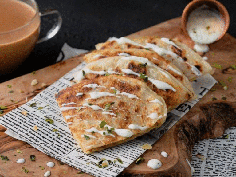 A wooden board holds several folded, golden-brown stuffed flatbreads drizzled with white sauce, garnished with herbs. A cup of chai and a small container of sauce sit nearby on the board, which is lined with newspaper.