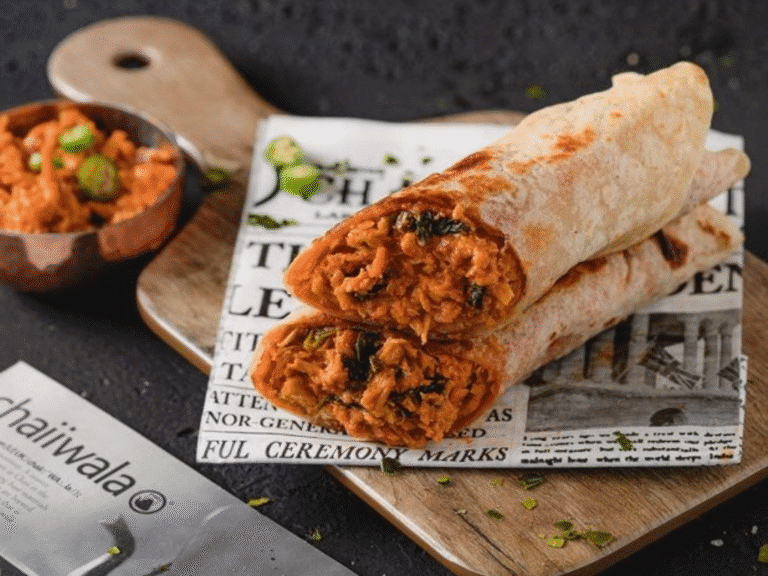 Two rolled flatbreads filled with a spicy, orange-colored filling are placed on newspaper-style parchment, served on a wooden board with a small bowl of extra filling garnished with herbs on the side.
