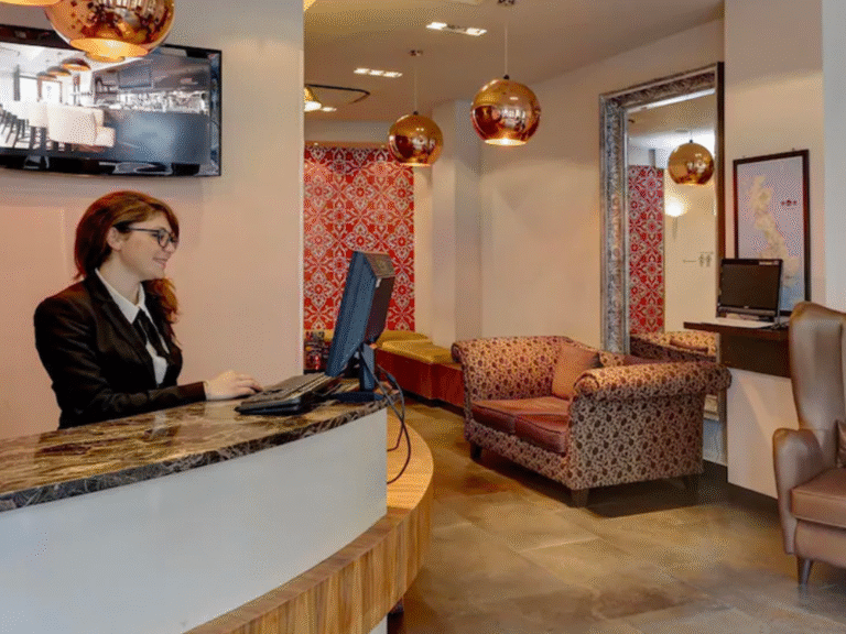 A woman in business attire works at a computer behind a marble reception desk in a stylish hotel lobby with patterned red walls, pendant lights, and comfortable seating.