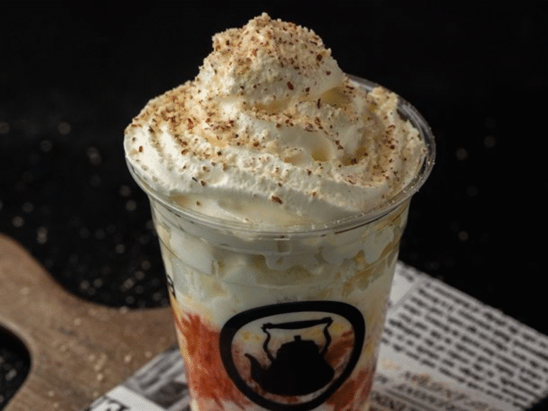 A plastic cup filled with a creamy dessert or drink, topped with whipped cream and sprinkled with brown crumbs, sits on a newspaper and wooden board against a dark background.