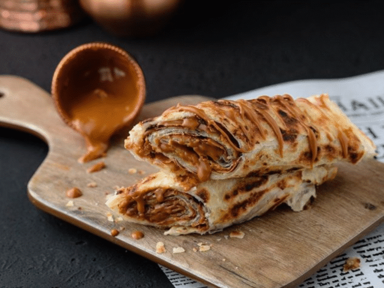 Two rolled flatbreads filled with caramel sauce are placed on a wooden serving board. A small cup, tipped over, spills caramel nearby. The background is dark with some newspaper under the board.