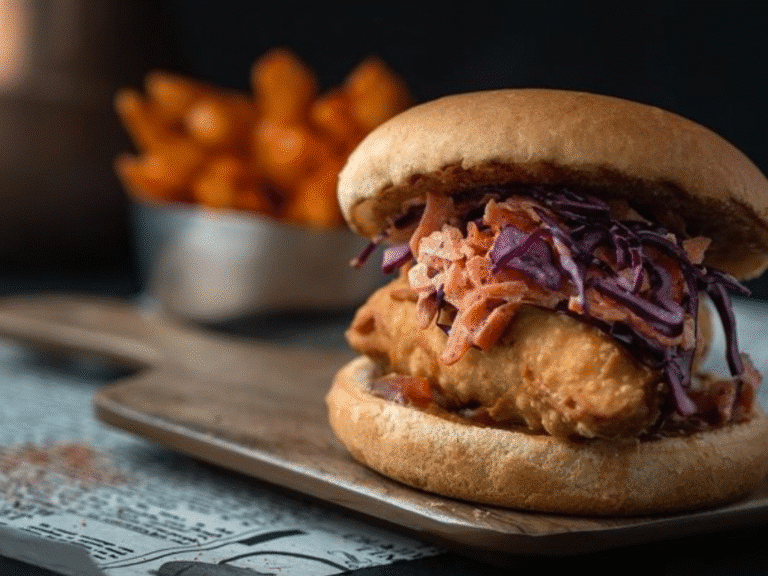 A crispy fried chicken sandwich topped with shredded purple and orange slaw is served on a wooden board, with a bowl of sweet potato fries blurred in the background.