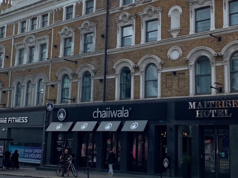 Street view of a building with arched windows; ground floor businesses include chaiiwala café with a black sign and white logo, a hotel entrance, and a fitness center. People and a bicycle are visible on the sidewalk.