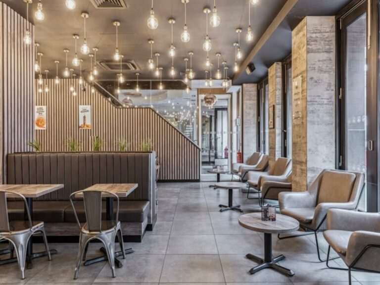 Modern café interior with grey armchairs, booth seating, wooden tables, tiled floor, and hanging pendant lights. Large windows line one side, letting in natural light. The overall design is stylish and contemporary.