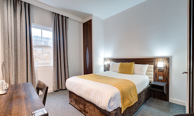 A modern hotel room with a double bed featuring white linens and a yellow throw blanket, a wooden headboard, bedside lights, a desk with a chair, and large windows with gray curtains.