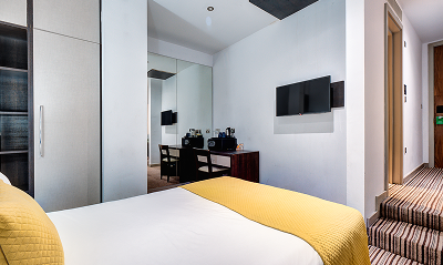 A modern hotel room with a bed featuring yellow bedding, a wall-mounted TV, a desk with a chair and coffee maker, mirrored closet doors, and striped carpet flooring.