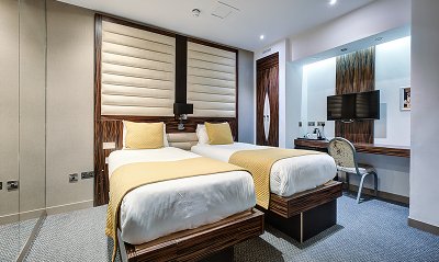 Modern hotel room with two single beds, yellow bedding, a TV mounted on the wall, a desk with a chair, and a large mirror. The decor is sleek with wood accents and soft lighting.