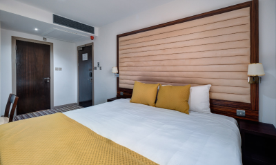 A modern hotel room with a large bed featuring yellow bedding and pillows, a padded headboard, bedside tables with lamps, a wooden door, and a striped carpet near the entrance.