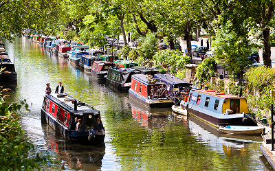 Colorful canal boats are moored along both sides of a leafy canal. A few people stand on one boat, and trees with green foliage overhang the water, creating a peaceful, vibrant scene.