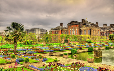 A vibrant formal garden with colorful flower beds, a pond, and neatly trimmed hedges sits in front of a historic brick mansion under a cloudy sky.