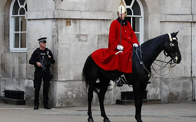 A mounted guard in a red coat and gold helmet sits on a black horse outside a historic stone building, while a police officer with a gun stands nearby.