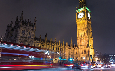 Nighttime view of Big Ben and the Palace of Westminster in London, with light trails from passing vehicles and a red double-decker bus visible in motion on the street in the foreground.