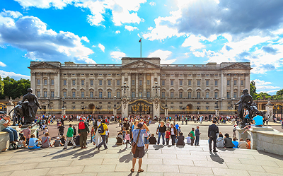 Crowds of people gather outside Buckingham Palace in London on a sunny day with blue skies and clouds. The historic building is prominent in the background, framed by statues and ornate gates at the front.