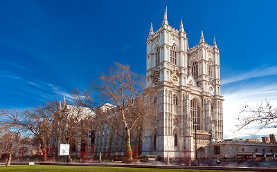A large Gothic-style cathedral with two tall towers stands against a bright blue sky, surrounded by bare trees and a green lawn.