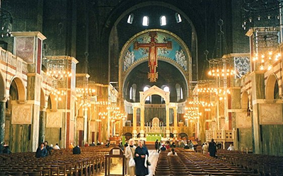 Interior of a grand cathedral with rows of wooden pews, ornate arches, chandeliers, and a large crucifix above the altar. Visitors are walking and sitting inside the softly lit, spacious church.