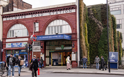 People walk past the entrance of Edgware Road Station in London, featuring a red-tiled facade and large arched windows. A green, plant-covered wall is visible to the right of the station entrance.