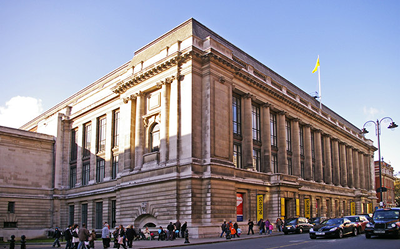 Large historic building with classical architecture, tall windows, columns, and a yellow flag on the roof. People walk along the sidewalk and enter through multiple doorways along the street. The sky is clear and blue.