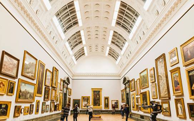 People walk through a spacious, bright art gallery with high arched ceilings, viewing numerous framed paintings displayed on the white walls. The room is well-lit with natural and artificial light.