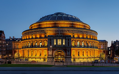 The Royal Albert Hall in London, illuminated with golden lights at dusk, displaying its circular architecture and grand entrance against a clear evening sky.