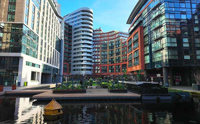 Modern glass and brick apartment buildings surround a landscaped courtyard with green plants, beside a calm canal with a small floating wooden structure under a bright blue sky.