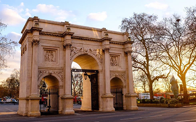 A stone arch monument with ornate carvings stands at a road intersection, surrounded by bare trees at sunrise. The sky is partly cloudy, and vehicles are visible in the background.