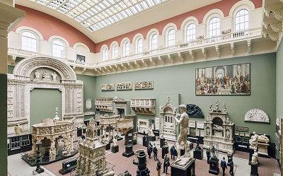 A spacious museum gallery with high ceilings, filled with classical sculptures, architectural models, and wall art. Visitors walk among the exhibits, and natural light streams through the glass roof.