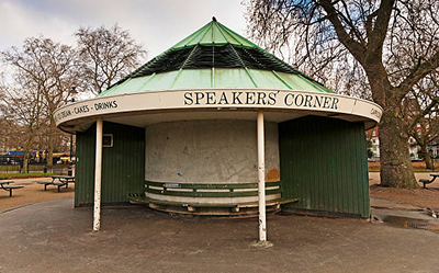 A small green and white round shelter labeled SPEAKERS CORNER stands in a park, surrounded by bare trees and empty benches on a cloudy day.