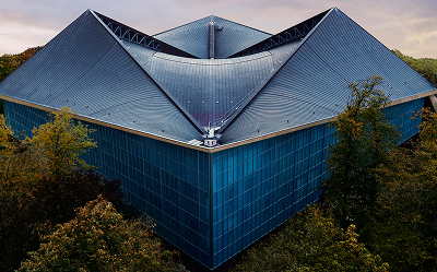 A modern building with a striking angular, folded roof, surrounded by trees. The structure features large blue-tinted glass walls and sharp geometric lines, creating a contemporary architectural look.