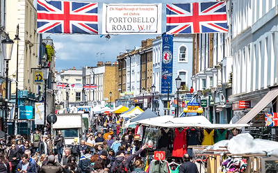 A bustling street market on Portobello Road in London, with Union Jack flags and a banner overhead, crowded with people and lined with colorful market stalls and historic buildings.