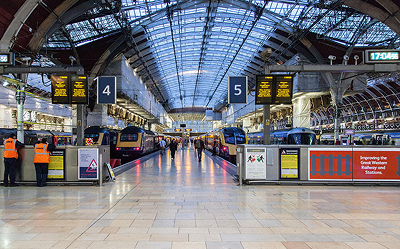 A brightly lit train station with a glass roof, platforms 4 and 5, electronic signs, two trains, and several people, including staff in orange vests, standing on the platform.
