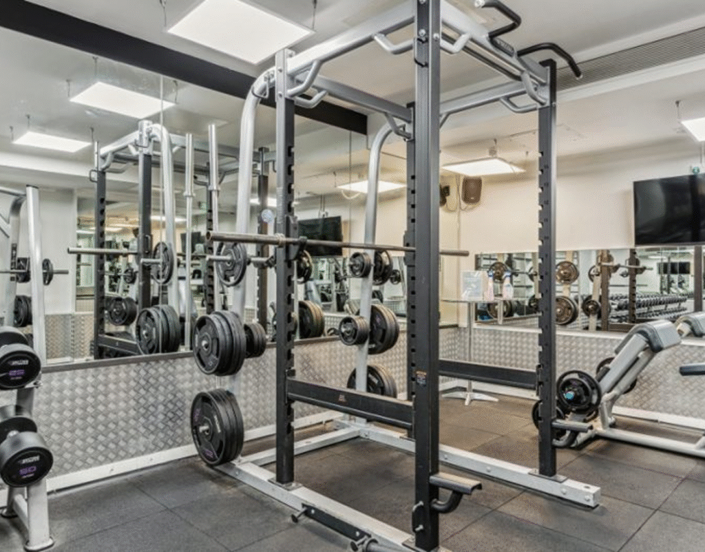 A gym with a power rack loaded with weights, mirrors on the walls, dumbbells on racks, and various exercise equipment including a bench and a leg press machine. The space is well-lit and has a modern, clean appearance.
