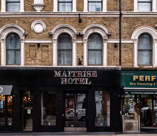 Street view of the Maitrise Hotel, featuring large arched windows on the upper floor, brick façade, and a neighboring dry cleaning shop with green signage.