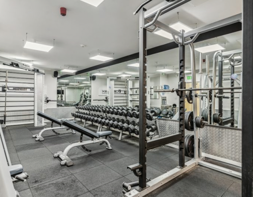 A modern gym with weight benches, free weights, dumbbells, a squat rack, and mirrors lining the walls. The space is well-lit with bright ceiling lights and has black rubber flooring.