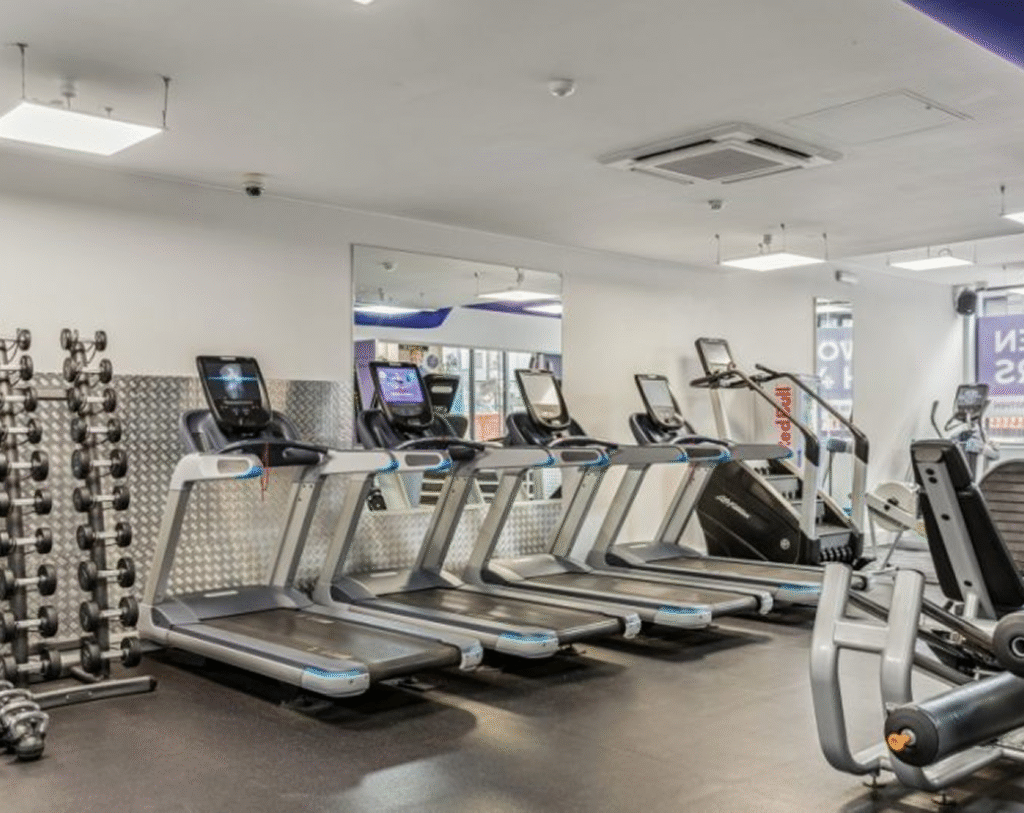 A modern gym with several treadmills, a row of dumbbells on a rack, exercise bikes, and large mirrors on the wall. The room is brightly lit with overhead lights.