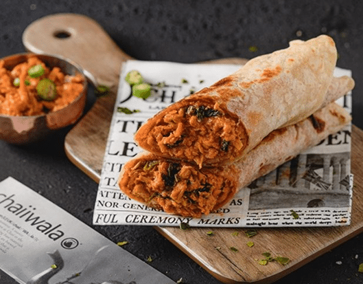 Two stuffed wraps filled with a spicy orange-colored filling are stacked on a wooden board lined with newspaper print. A small bowl of chutney garnished with sliced chilies is beside them, against a dark background.